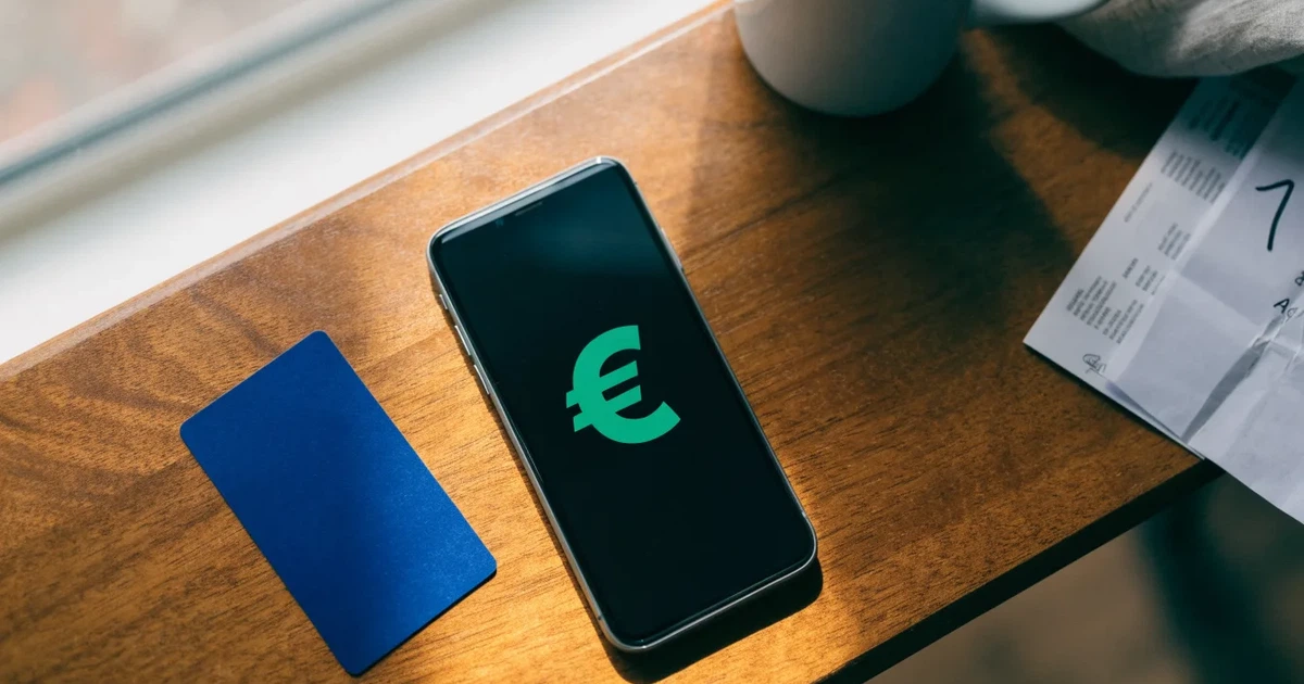 Um telemóvel e um cartão bancário lado a lado numa mesa de madeira, com algumas notas de euro ao lado